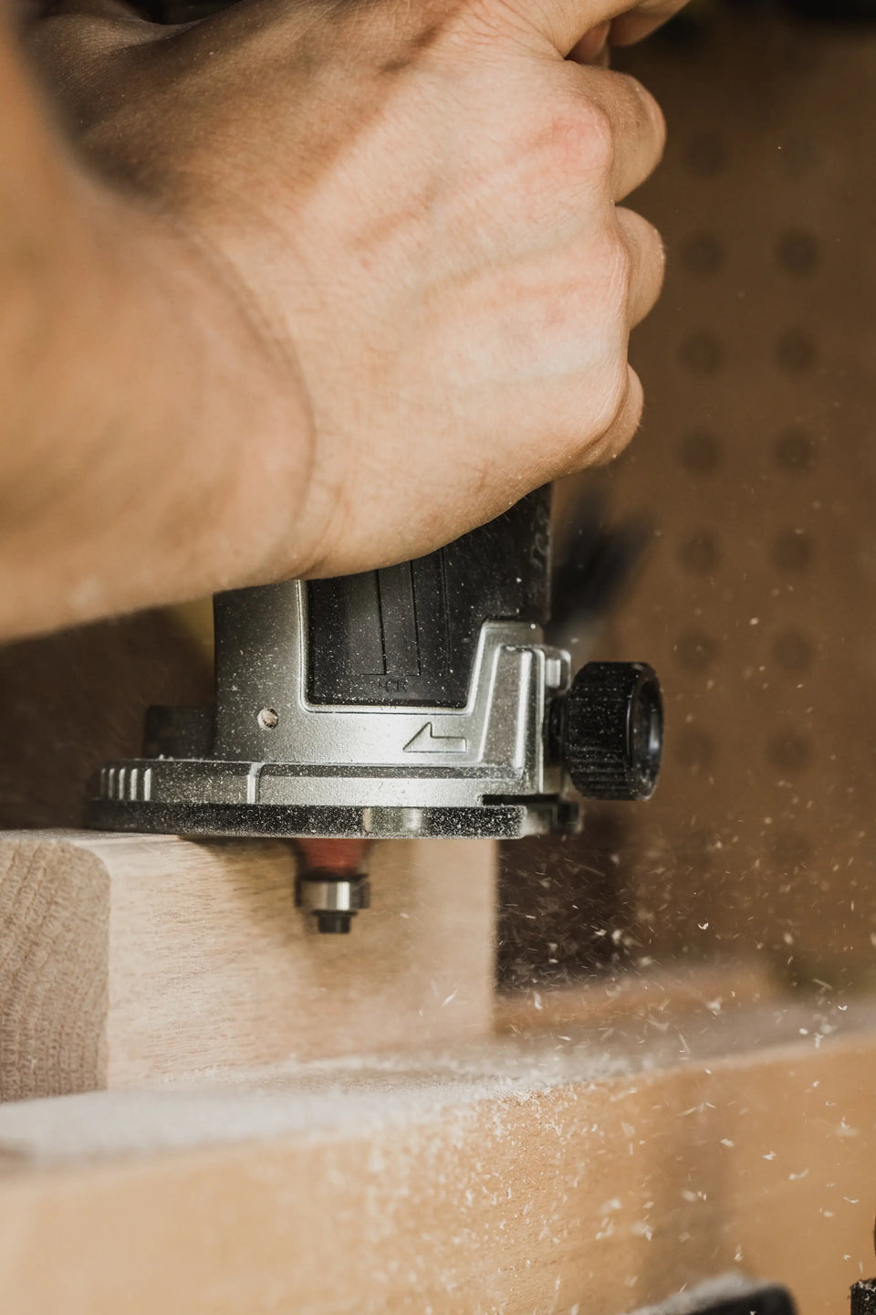 Close-up of a hand using an electric router on wood, creating sawdust, in a workshop setting.